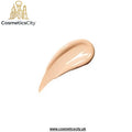 A swatch of liquid foundation or concealer in a warm beige shade, shown as a glossy smear on a plain white background with the CosmeticsCity logo in the top left and website text at the bottom.
